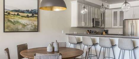 A modern kitchen with a central island, pendant lighting, and a dining area with chairs and a table.