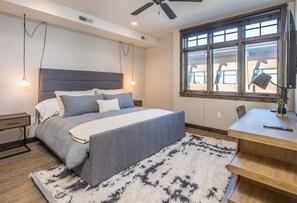 Ein Schlafzimmer mit einem Bett, Nachttischen, einem Schreibtisch, einem Stuhl, einem Deckenventilator und einem großen Fenster.