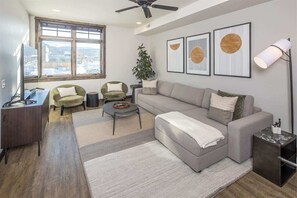 A modern living room with a grey sofa, a small round coffee table, a desk with a computer, and a large window offering a city view.