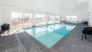 A modern indoor swimming pool with clear glass walls, a tiled floor, and black chairs around the pool. - Luxury Home w/ Indoor Pool & Slide, Sports Court, Sleeps 40 (Garden City)