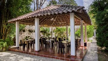 A covered outdoor dining area with tables and chairs, surrounded by greenery and trees.