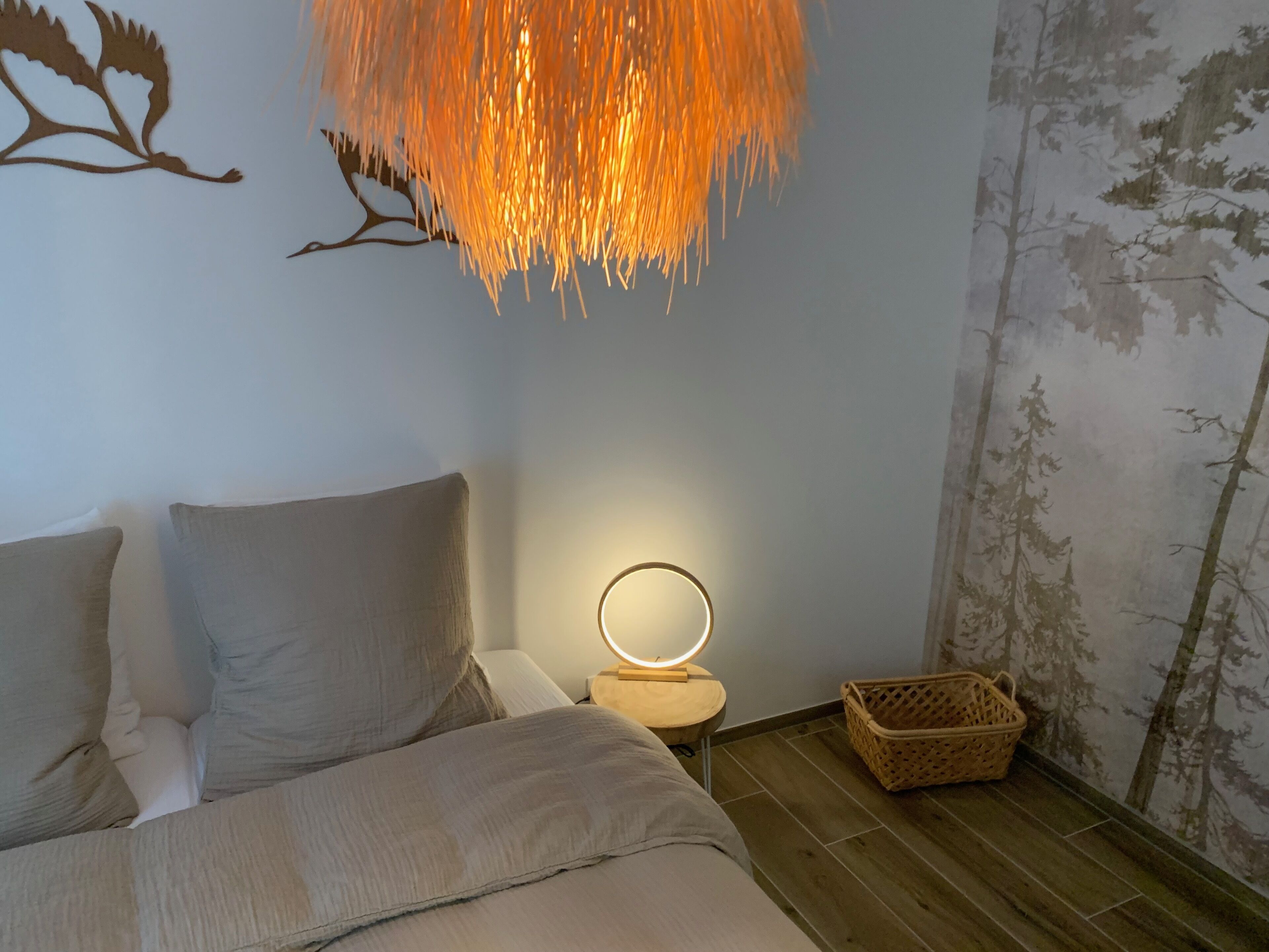 Een slaapkamer met een bed, een nachtkastje met een lamp, een muur met een boomdecoratie en een houten vloer.