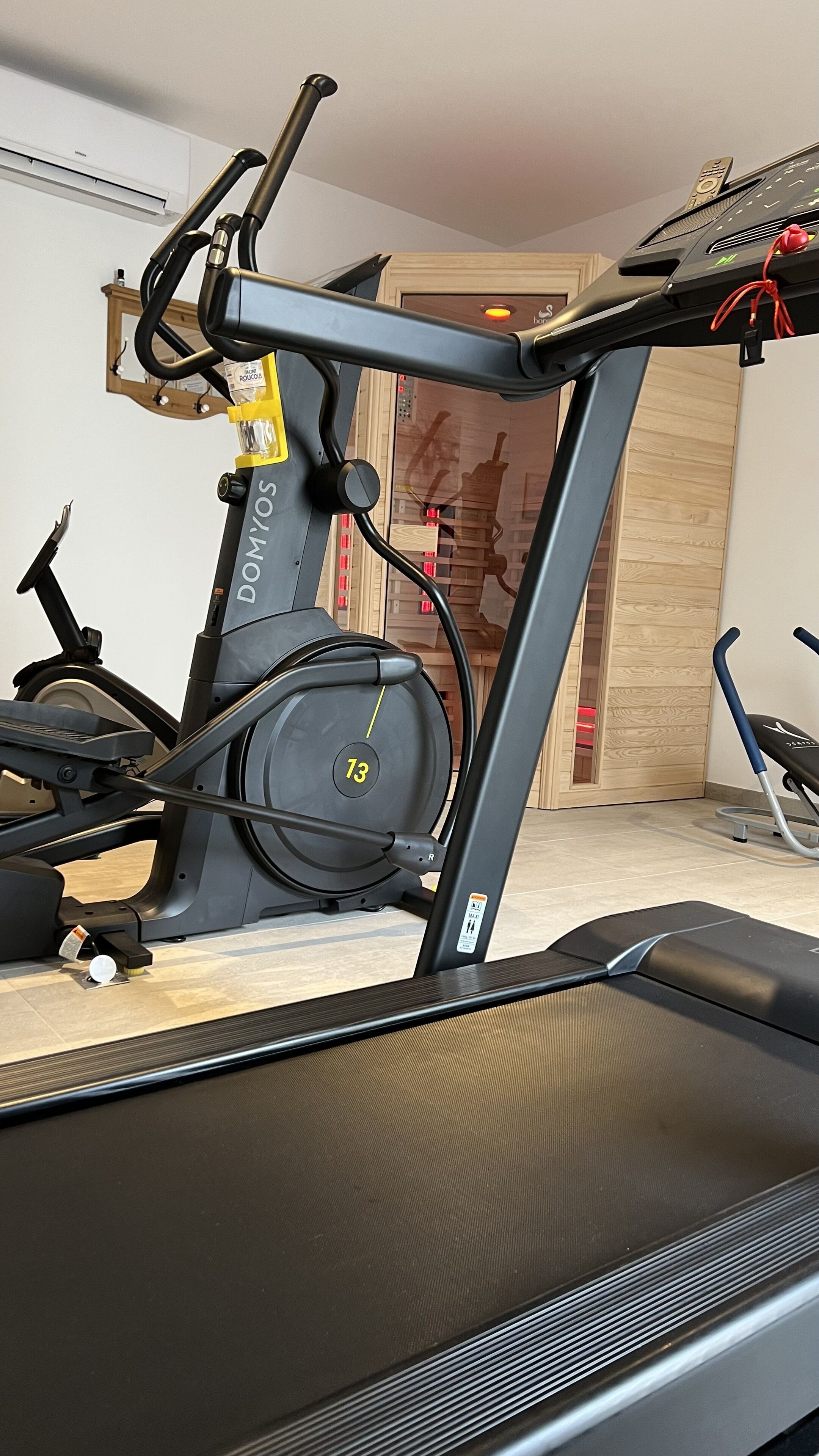 A stationary bike with a digital display, a treadmill, and exercise equipment in a room with wooden flooring and a mirror.