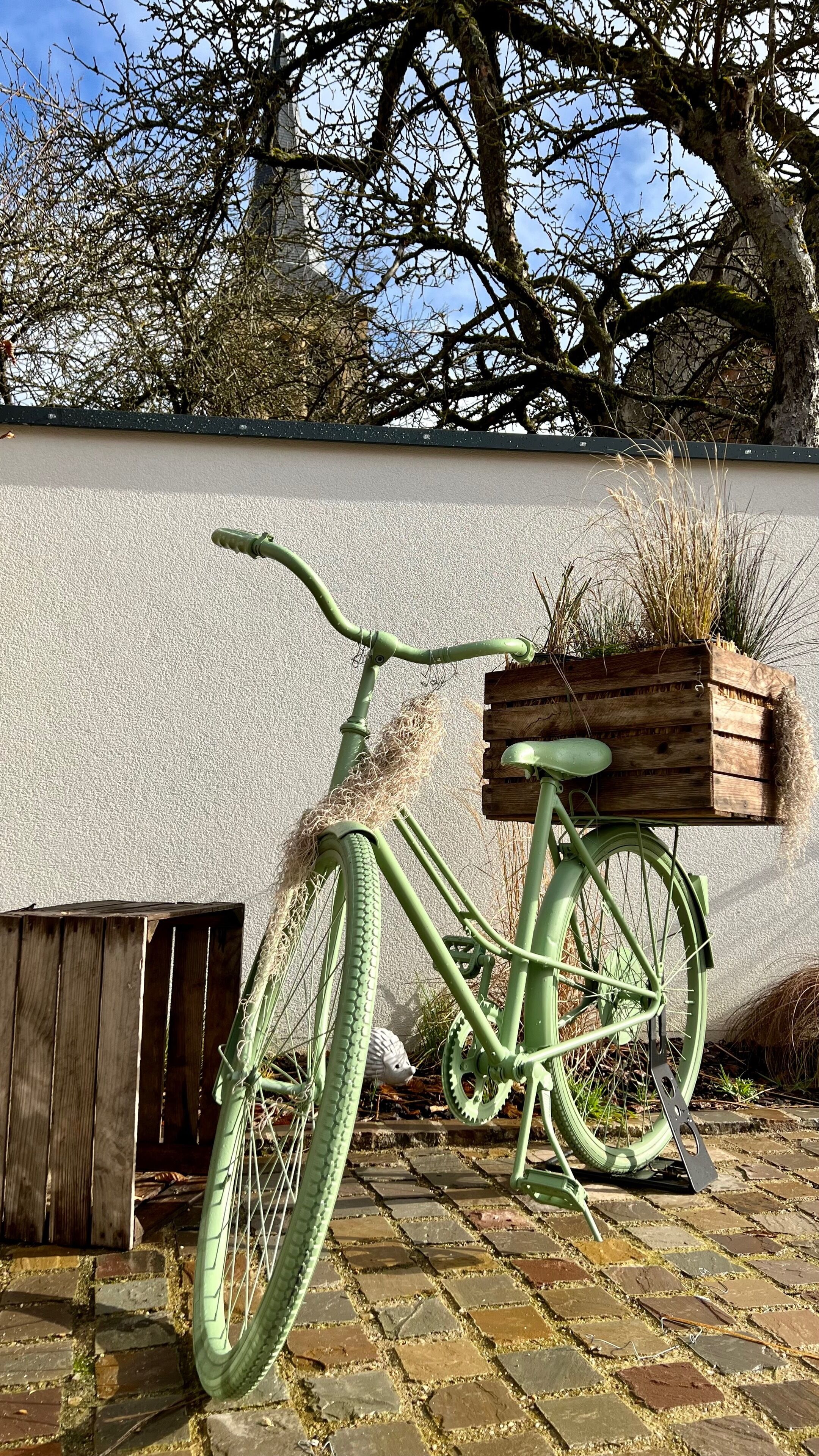 Un vélo vert d’époque, doté d’un panier en bois fixé à l’arrière, est garé sur une surface pavée de pierres, adossé à un mur blanc.