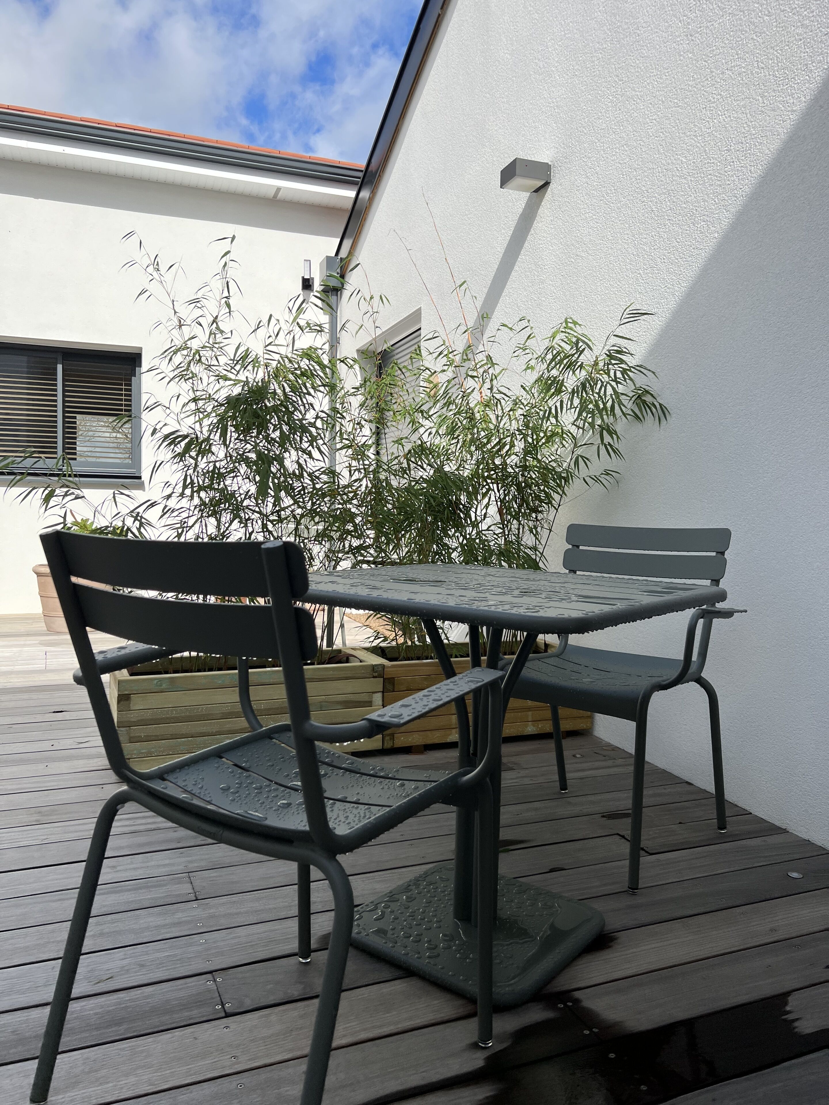 A patio with a table and chairs, a wooden deck, a white wall, and a bamboo plant.