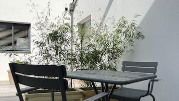A patio with a table and chairs, a wooden deck, a white wall, and a bamboo plant.