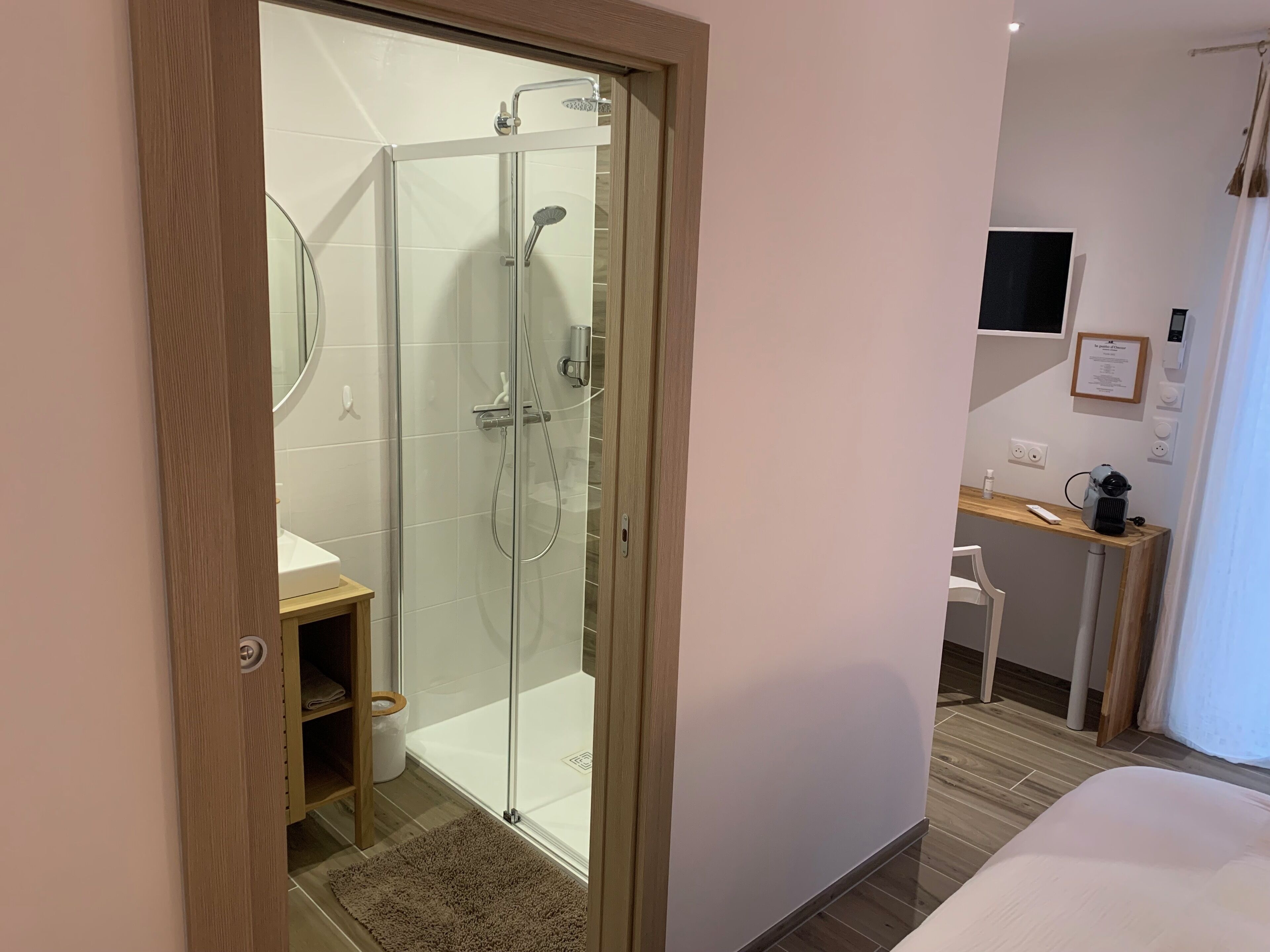 A modern hotel bathroom with a glass-enclosed shower, a wooden vanity, and a small shelf.