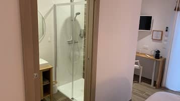A modern hotel bathroom with a glass-enclosed shower, a wooden vanity, and a small shelf.
