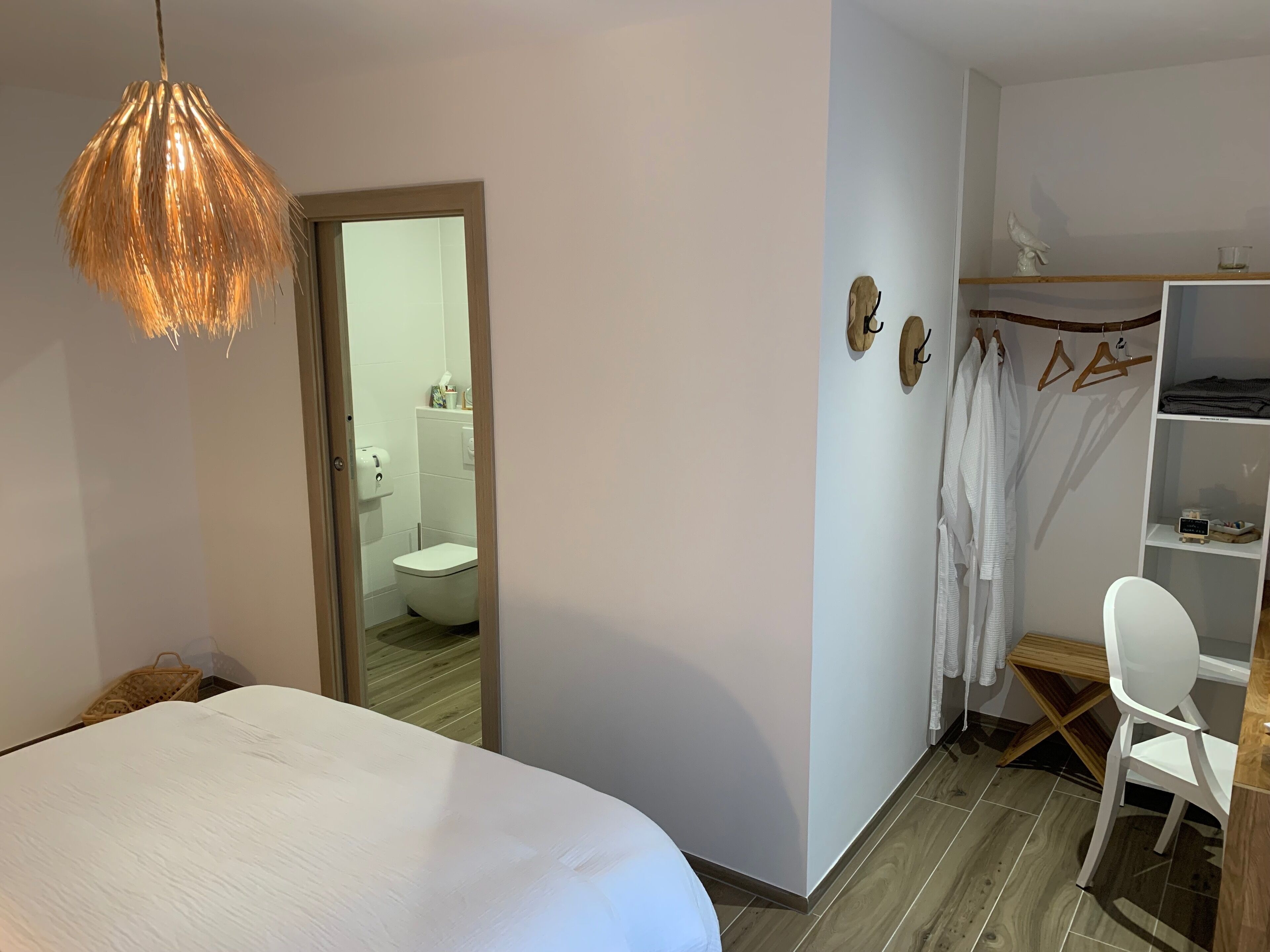 A bedroom with a bed, a hanging light fixture, a wooden floor, a white wardrobe, a white chair, and a bathroom visible through an open door.