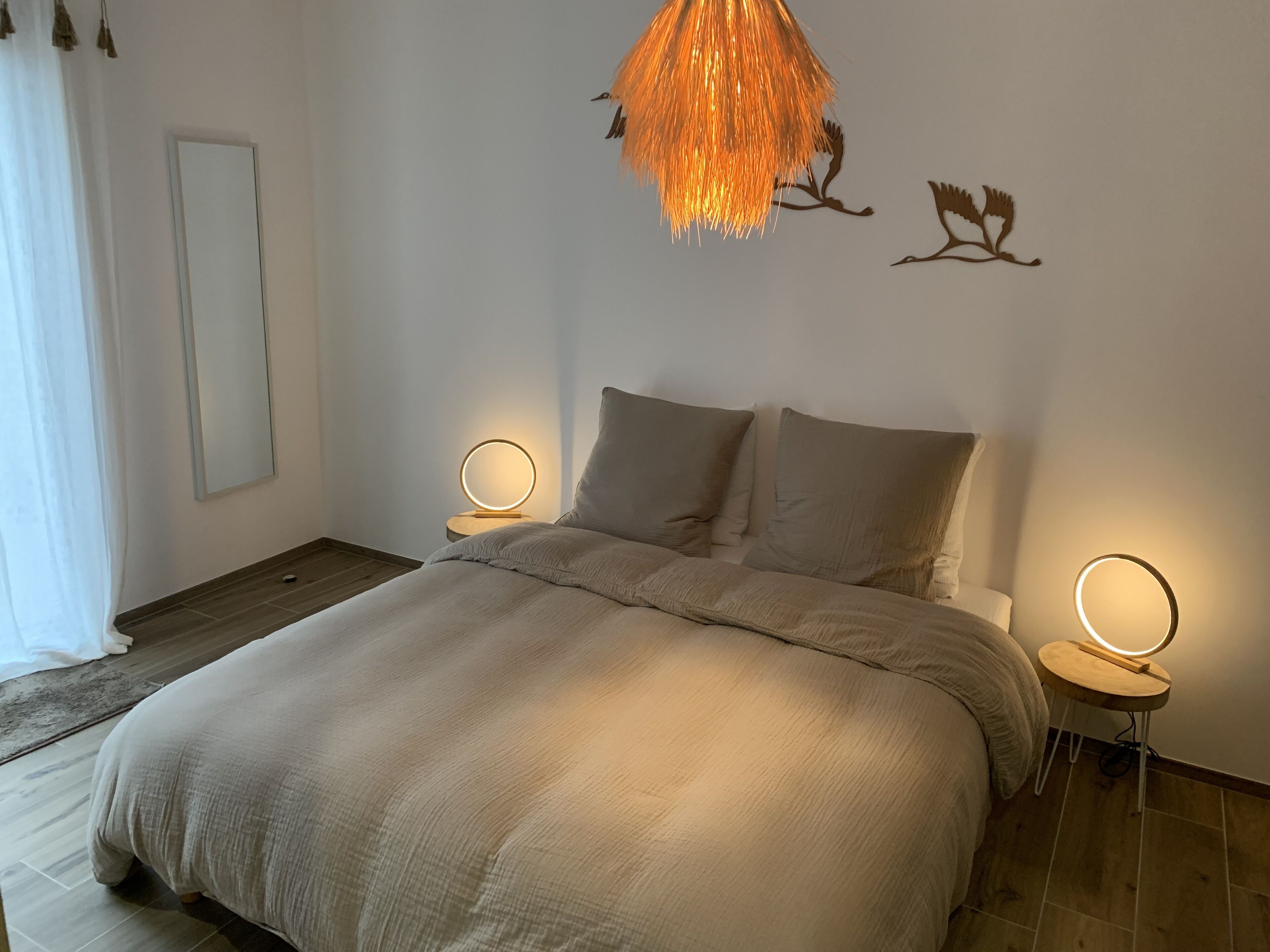 Un dormitorio con una cama grande, dos mesitas de noche con espejos redondos, una lámpara colgante y cuadros en la pared.
