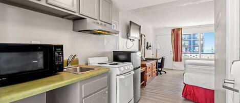 A compact kitchen with a microwave, stove, and sink. A bed with a red skirt is visible in the background.