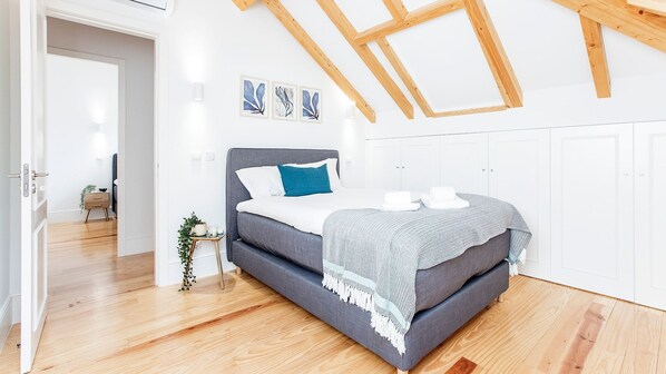Une chambre moderne avec un canapé-lit gris, une table de chevet blanche et une petite plante. Trois œuvres d’art encadrées sont accrochées au mur au-dessus du lit.