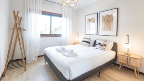 Moderni hotellihuone, jossa on sänky, puinen maalausteline ja kaksi kehystettyä taideteosta seinällä.