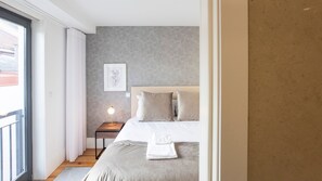 A modern bedroom with a large bed, a bedside table with a lamp, a framed picture on the wall, and a view through a window.