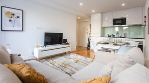 A modern living room with a flat-screen TV, a dining area with a glass-top table, and a kitchen with a microwave.
