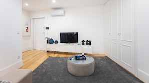 A modern living room with a flat-screen TV, a white console table with decorative items, a grey ottoman, and a grey carpet.