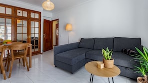 A modern living room with a grey sectional sofa, wooden dining table with chairs, a small round wooden table with a plant, and a large window with a view of the outside.