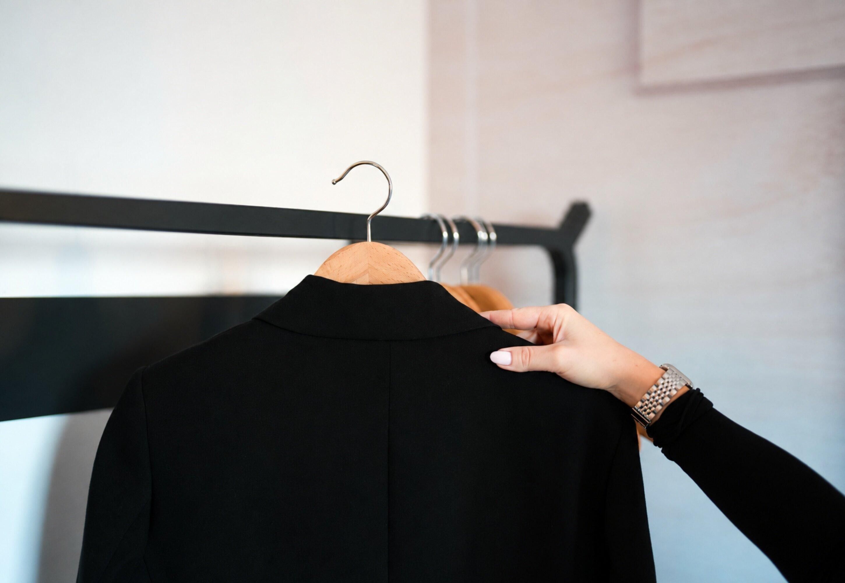 A hand holding a black garment on a wooden hanger against a wall with a black horizontal bar.