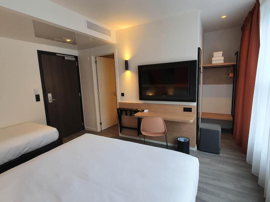 A hotel room with a bed, a desk, a chair, a TV, a wardrobe, and a trash bin.