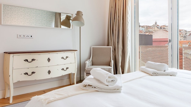 Habitació d'hotel amb un llit, una petarrera, una cadira, una làmpada i vista a edificis a través d'una finestra.