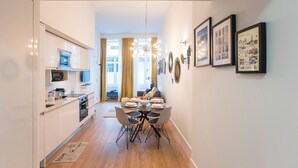 Une cuisine moderne avec des meubles blancs, un four à micro-ondes et un coin repas comprenant une table et des chaises en verre. Des tableaux encadrés ornent le mur, et un petit sapin de Noël est installé dans un coin.
