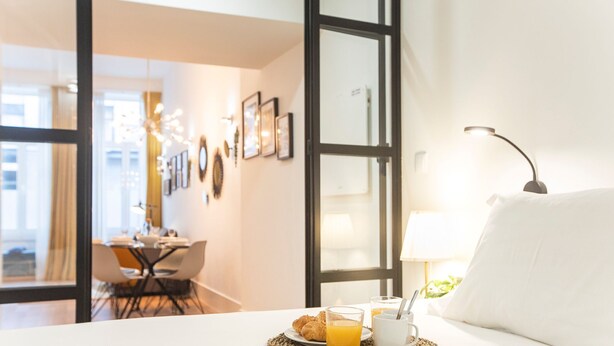 Une chambre d’hôtel avec un lit, un plateau de petit-déjeuner comprenant des croissants et du jus d’orange, et un coin repas visible à travers une porte ouverte.