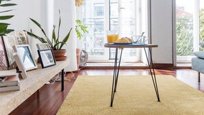 A cozy living room with a wooden floor, a round table with a yellow rug, a potted plant, and a window with a view of buildings. - Bonjardim Studio w/ Balcony by LovelyStay (Porto)