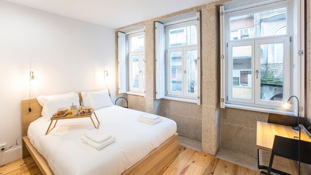 Habitación de hotel con cama, mesita de noche y vistas a la ciudad a través de amplios ventanales.