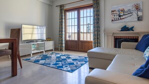 Een woonkamer met een bank, een televisie, een open haard en een groot raam met gordijnen.