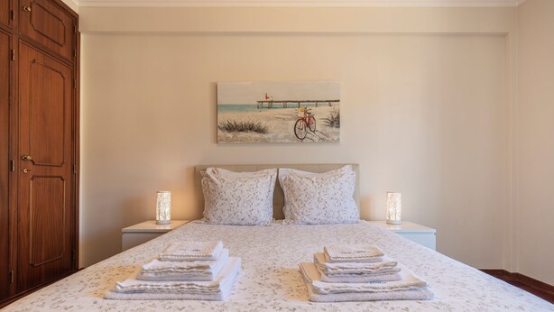 Een slaapkamer met een bed, twee nachtkastjes, een houten garderobe en een schilderij met een strandthema aan de muur.