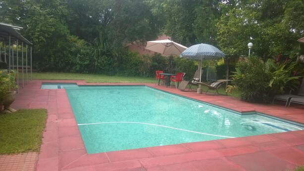 A rectangular swimming pool with a tiled edge, surrounded by a red brick patio. There are two red chairs and a table with a patterned umbrella nearby. - Gloria's Bed And Breakfast Accommodation (Livingstone)