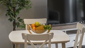 Een eetgedeelte met een witte tafel, stoelen, een mand met fruit, een bloempot en een hanglamp.