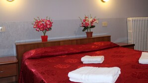A hotel room with a red bedspread, two potted plants, a wooden nightstand, and a radiator. - Double Room (Roma)
