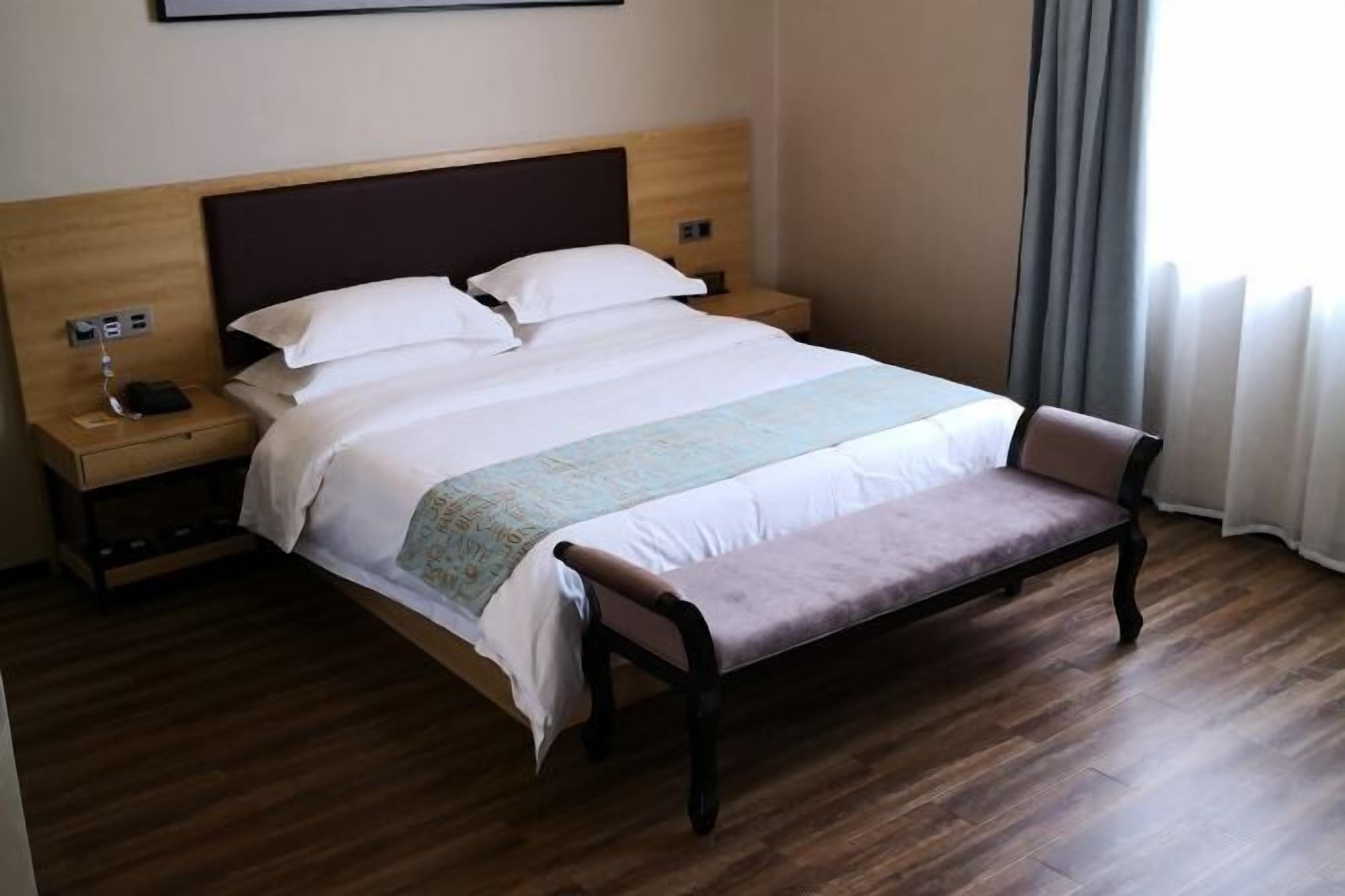 A hotel room with a wooden bed, a bench, a nightstand with a phone, and a window with curtains.