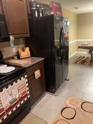 Una cocina con un refrigerador negro, un microondas y una estufa. Hay una tabla de cortar de madera y un par de tijeras sobre la encimera.