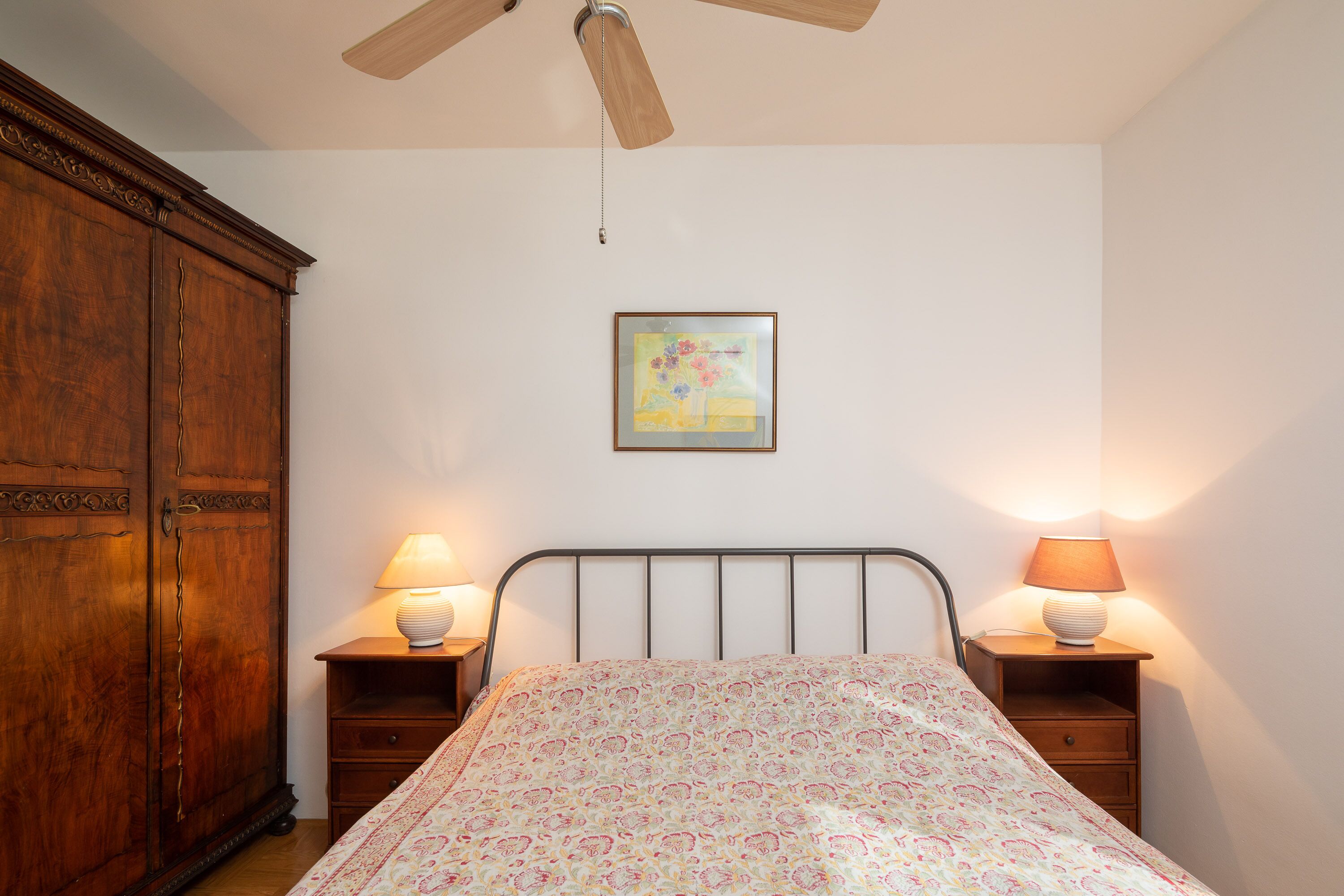 A bedroom with a bed, wooden wardrobe, two bedside tables with lamps, a ceiling fan, and a framed picture on the wall.