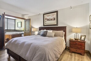 Un dormitorio con una cama grande, cabecera de madera, mesitas de noche con lámparas, un cuadro enmarcado en la pared y una ventana con persianas.