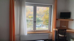 A hotel room with a bed, a desk, a chair, a TV, and a window with a view of trees and a body of water. - Hotel Gasthof zum Ritter (Hassmersheim)