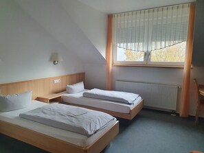A hotel room with two single beds, a nightstand, a lamp, a window with blinds, and a radiator.
