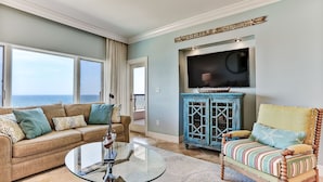 A living room with a large window, a sofa, a chair, a glass coffee table, and a TV mounted on the wall. - Sensational Sandcastles Condo/Oceanfront-Amelia (Fernandina Beach)