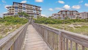 A wooden boardwalk leading to a multi-story hotel with balconies, surrounded by greenery and sand dunes. - Sensational Sandcastles Condo/Oceanfront-Amelia (Fernandina Beach)