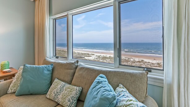 A room with a large window overlooking a beach, a sofa with patterned cushions, and a small table with a green container. - Sensational Sandcastles Condo/Oceanfront-Amelia (Fernandina Beach)