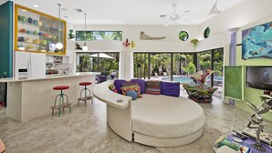 A modern living room with a curved sofa, a kitchen with a glass display cabinet, and a large window overlooking a pool.