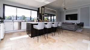 Una cocina moderna con una isla grande, taburetes de bar y un sofá gris en un amplio salón con un gran ventanal y un ventilador de techo.