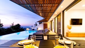 Un área de comedor exterior moderna con piscina, asientos cómodos e interior bien iluminado.
