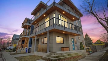 A modern multi-story residential building with a balcony, large windows, and a well-lit entrance.