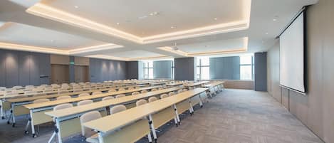 Uma sala de conferências com mesas dispostas em fileiras, uma tela de projetor e grandes janelas.