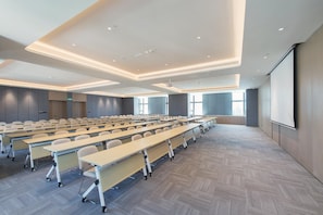 Uma sala de conferências com mesas dispostas em fileiras, uma tela de projetor e grandes janelas.