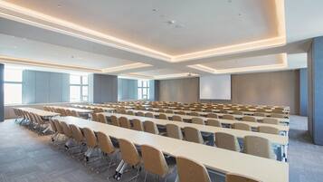 Een vergaderzaal met rijen tafels en stoelen, grote ramen en een projectiescherm.