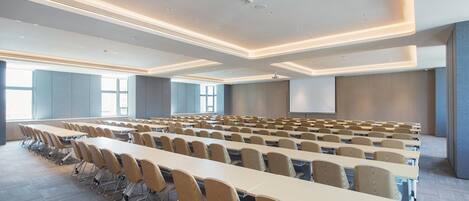 A conference room with rows of tables and chairs, large windows, and a projection screen.